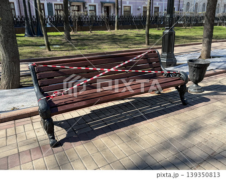 Park bench wrapped with warning tape in urban area with pavement, trees in daylight scene. Public safety, restriction concept, urban environment, city management, outdoor infrastructure 139350813