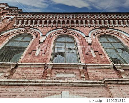 The old red brick industrial building features arched windows and decorative masonry. It stands tall against a clear blue sky, showcasing its unique architecture from a low angle. The old red brick industrial building features arched windows and decorative masonry. It stands tall against a clear blue sky, showcasing its unique architecture from a low angle. 139351211