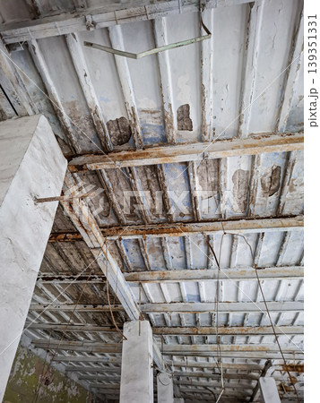 A ceiling with peeling white paint and rusted metal beams in an abandoned interior. The structure displays clear signs of age and neglect. The environment appears desolate. A ceiling with peeling white paint and rusted metal beams in an abandoned interior. The structure displays clear signs of age and neglect. The environment appears desolate. 139351331