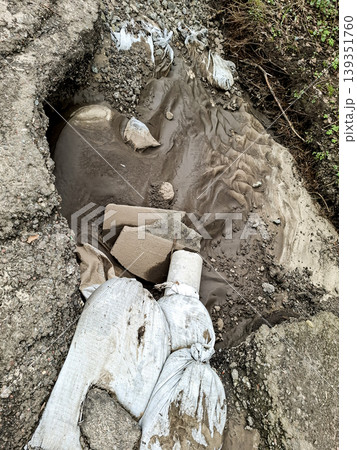 A large pothole on an asphalt road fills with wet mud and debris. Workers use sandbags for temporary repair and control of erosion in the area. The scene shows ongoing roadway maintenance. A large pothole on an asphalt road fills with wet mud and debris. Workers use sandbags for temporary repair and control of erosion in the area. The scene shows ongoing roadway maintenance. 139351760