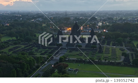 An enchanting evening aerial shot of the Prambanan Hindu Temple complex in Indonesia, featuring its iconic towering stone spires illuminated against a misty twilight sky and a sprawling cityscape. 139352742