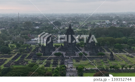 An enchanting evening aerial shot of the Prambanan Hindu Temple complex in Indonesia, featuring its iconic towering stone spires illuminated against a misty twilight sky and a sprawling cityscape. 139352744