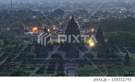 An enchanting evening aerial shot of the Prambanan Hindu Temple complex in Indonesia, featuring its iconic towering stone spires illuminated against a misty twilight sky and a sprawling cityscape. 139352745