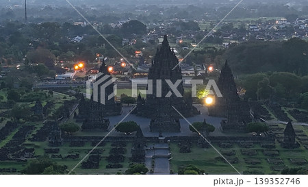 An enchanting evening aerial shot of the Prambanan Hindu Temple complex in Indonesia, featuring its iconic towering stone spires illuminated against a misty twilight sky and a sprawling cityscape. 139352746