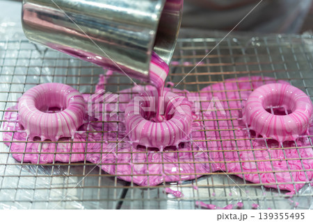 Fresh donuts being coated with pink glaze on wire rack showing baking process glossy texture and sweet dessert preparation in kitchen Fresh donuts being coated with pink glaze on wire rack showing baking process glossy texture and sweet dessert preparation in kitchen 139355495