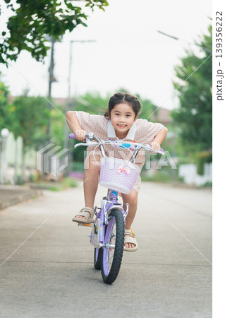 Happy little girl joyfully riding her bicycle on a sunny day, showcasing the thrill of childhood adventures and the beauty of outdoor playtime in a neighborhood setting. 139356222