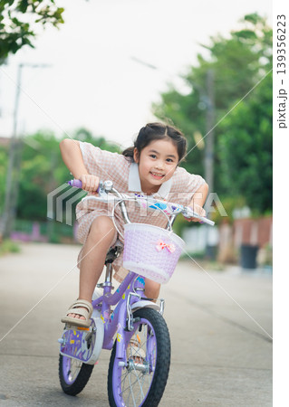 Joyful Young Girl Riding Bicycle with Purple Basket on Sunny Street, Embracing Freedom and Fun in Childhood Adventure 139356223