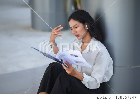 Overworked businesswoman sits by a column reviewing financial documents on a clipboard, gesturing with concern in a modern office space for stress, planning, and workload concepts. Overworked businesswoman sits by a column reviewing financial documents on a clipboard, gesturing with concern in a modern office space for stress, planning, and workload concepts. 139357547