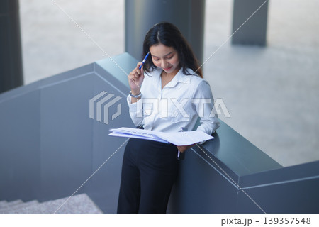 Professional businesswoman leans on an indoor railing, reviewing financial documents with a pen in a modern office, showing corporate planning, business analysis, productivity, and focused work. Professional businesswoman leans on an indoor railing, reviewing financial documents with a pen in a modern office, showing corporate planning, business analysis, productivity, and focused work. 139357548