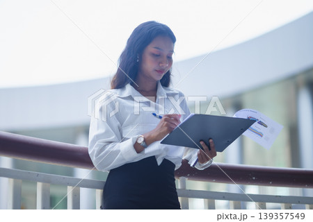 Professional businesswoman stands near a railing and writes on a clipboard with financial charts in a bright office atrium, showing planning, report review, and corporate workflow tasks. Professional businesswoman stands near a railing and writes on a clipboard with financial charts in a bright office atrium, showing planning, report review, and corporate workflow tasks. 139357549
