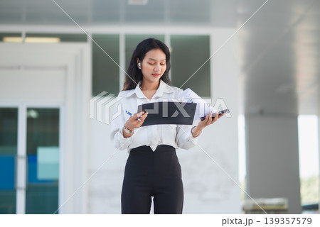 Businesswoman reviews charts on a clipboard and uses a smartphone in a modern office lobby, showing financial analysis, multitasking, digital communication, and office work planning. 139357579