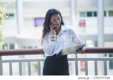 Businesswoman talks on a smartphone while holding a clipboard at an office railing, combining mobile communication, financial paperwork, corporate planning, and productivity for business workflow. Businesswoman talks on a smartphone while holding a clipboard at an office railing, combining mobile communication, financial paperwork, corporate planning, and productivity for business workflow. 139357580