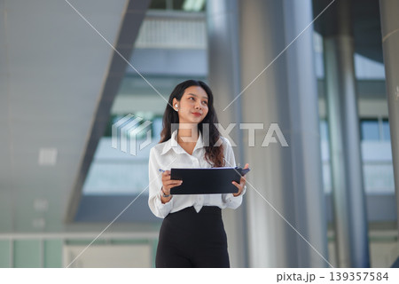 Confident businesswoman holds a clipboard and pen while looking away in a modern office atrium, ideal for corporate strategy, project planning, leadership, and business career growth. 139357584