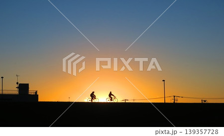夕陽を浴びながら土手を自転車で直進する二人 夕陽を浴びながら土手を自転車で直進する二人 139357728