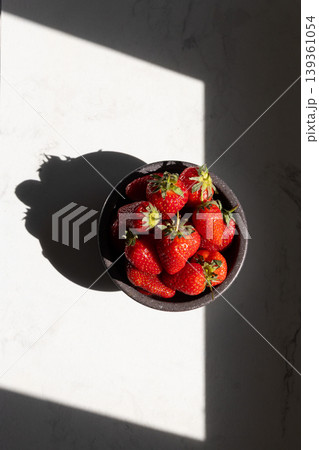 Fresh red strawberries in black bowl on white marble surface. Sunlight casting dramatic shadows. Healthy summer fruit and natural food concept for recipes and nutrition. Fresh red strawberries in black bowl on white marble surface. Sunlight casting dramatic shadows. Healthy summer fruit and natural food concept for recipes and nutrition. 139361054