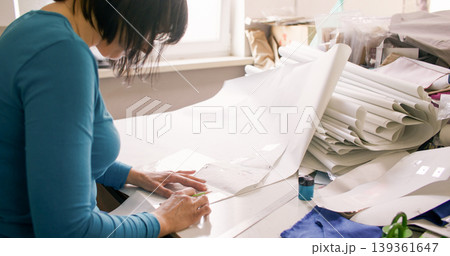 Dressmaking process. Tailor desk. Woman hand creating garment patterns with ruler making notes on table cluttered with paper drafts. 139361647