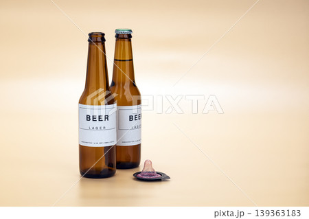 Two brown craft lager beer bottles placed beside an unwrapped pink condom on a warm beige studio background, symbolizing responsible safe sex and alcohol awareness Two brown craft lager beer bottles placed beside an unwrapped pink condom on a warm beige studio background, symbolizing responsible safe sex and alcohol awareness 139363183