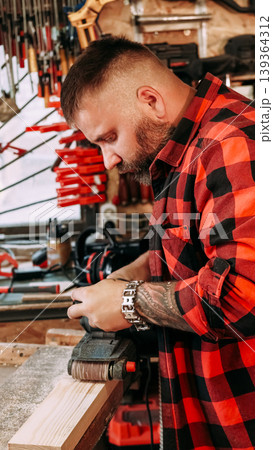 Sanding wood. Carpentry tool. Joiner man smoothing plank surface with professional belt sander equipment in garage workshop interior. Sanding wood. Carpentry tool. Joiner man smoothing plank surface with professional belt sander equipment in garage workshop interior. 139364312