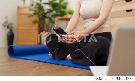 Asian women sitting on yoga mat using smartphone mobile phone app for fitness workout exercise training at home living room healthy lifestyle wellness 139365378