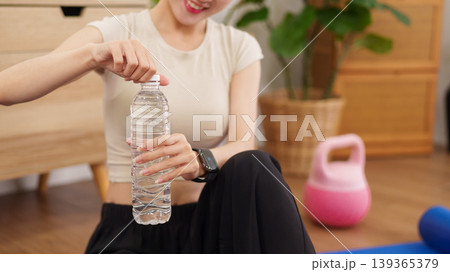 Asian women opening water bottle after home workout exercise session sitting floor with kettlebell fitness equipment healthy lifestyle wellness hydration 139365379
