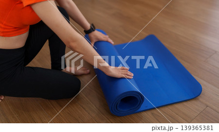 Asian women rolling blue yoga mat preparing fitness workout exercise training session on wooden floor at home gym studio wearing orange shirt black pants Asian women rolling blue yoga mat preparing fitness workout exercise training session on wooden floor at home gym studio wearing orange shirt black pants 139365381