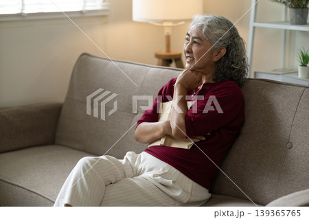 Asian senior woman relaxing reading book at home living room sofa thoughtful expression enjoying quiet leisure moment cozy domestic comfort daily life 139365765