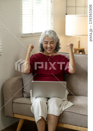 Asian senior woman celebrating online business success with joyful fist pump gesture at home achieving victory remote work lifestyle Asian senior woman celebrating online business success with joyful fist pump gesture at home achieving victory remote work lifestyle 139365806