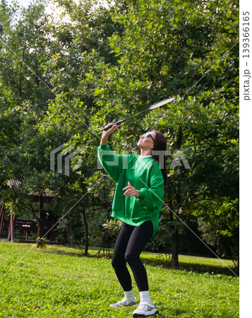 Young woman playing badminton in green park. Athletic adult holding racket during outdoor sports activity. Summer leisure, active lifestyle and healthy recreation in nature concept. 139366165