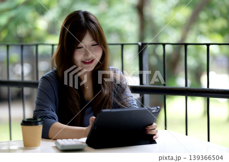 Young Asian woman performs digital tasks utilizing technology in a relaxing outdoor cafe environment fostering productivity and learning Young Asian woman performs digital tasks utilizing technology in a relaxing outdoor cafe environment fostering productivity and learning 139366504