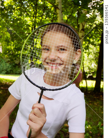 Young girl peeking through badminton racket in sunny park. Smiling child holding sports gear with green trees and outdoor nature background Young girl peeking through badminton racket in sunny park. Smiling child holding sports gear with green trees and outdoor nature background 139366541