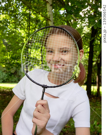 Young girl peeking through badminton racket in sunny park. Smiling child holding sports gear with green trees and outdoor nature background 139366542