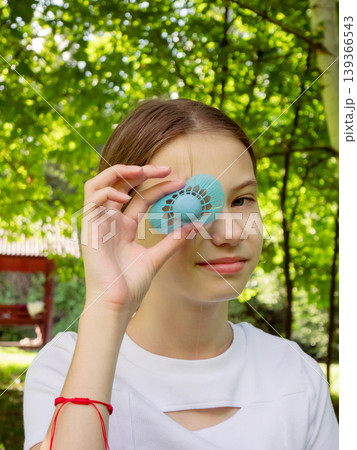 Girl holding blue shuttlecock in front of eye in sunny park. Teenager playing outdoor sport game. Summer leisure activity and recreation concept. Young woman enjoying sunny day outside. 139366543