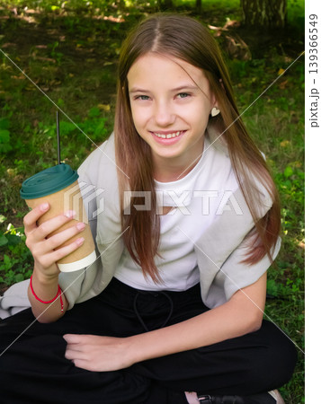 Young girl smiling with disposable coffee cup in lush green park. Casual outdoor portrait of happy teen girl relaxing on grass amid nature Young girl smiling with disposable coffee cup in lush green park. Casual outdoor portrait of happy teen girl relaxing on grass amid nature 139366549