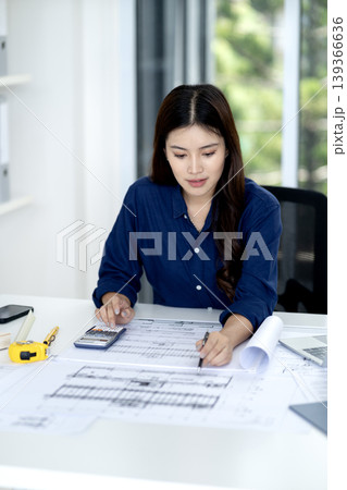 Focused Asian architect reviews building designs making precise calculations on intricate plans at contemporary office workspace Focused Asian architect reviews building designs making precise calculations on intricate plans at contemporary office workspace 139366636