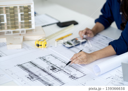 Asian adult architect professional carefully analyzes detailed building blueprints using calculator at office table Asian adult architect professional carefully analyzes detailed building blueprints using calculator at office table 139366638