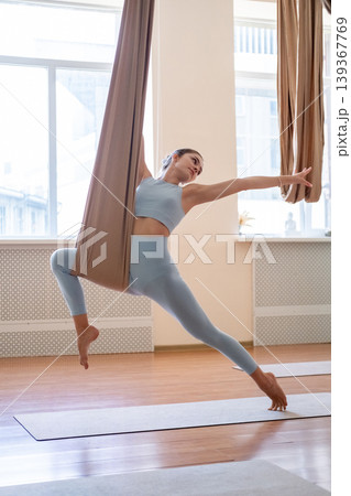 Fly yoga. Woman stretching. Young athlete doing gymnastics exercise in leaning pose supported by fabric hammock wrapped around leg in gym. 139367769