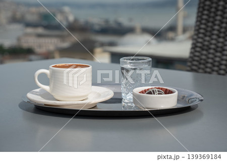 Turkish coffee in white cup on tray with water glass and chocolate dessert on table with blurred cityscape and Bosphorus background. Travel lifestyle, hospitality, urban leisure, beverage 139369184