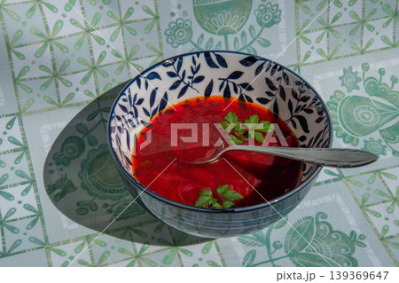 Ukrainian beetroot soup borscht poured into floral bowl over green cloth. Authentic borsch looks bright under sunlight with green herbs and metal spoon. Ukrainian beetroot soup borscht poured into floral bowl over green cloth. Authentic borsch looks bright under sunlight with green herbs and metal spoon. 139369647