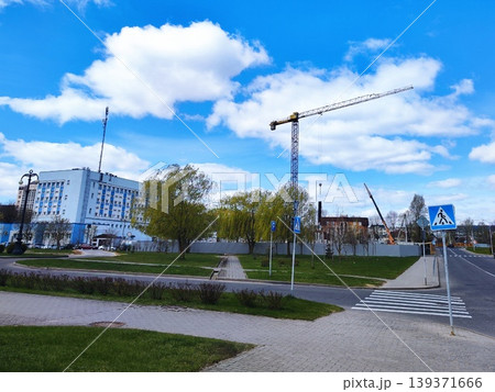 Street construction scene with a visible crane and ongoing development behind fencing. A large building stands nearby, characterized by white and blue colors Street construction scene with a visible crane and ongoing development behind fencing. A large building stands nearby, characterized by white and blue colors 139371666