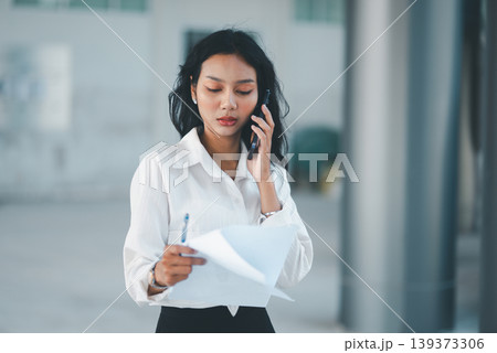 Professional businesswoman talking on smartphone while reviewing documents and holding a pen in a modern office, ideal for business communication, multitasking, client support, and workflow 139373306