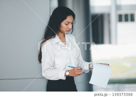 Professional woman checking paperwork with pen while standing in a modern office, suitable for corporate administration, report review, business planning, concentration, and workplace routine. Professional woman checking paperwork with pen while standing in a modern office, suitable for corporate administration, report review, business planning, concentration, and workplace routine. 139373308