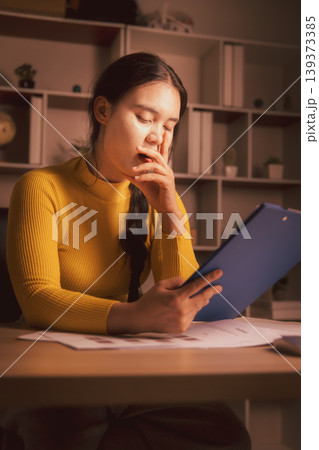 Sleepy young Asian woman yawning while reviewing clipboard documents in cozy home office at night, illustrating paperwork fatigue, overtime stress, deadline pressure and evening workload. 139373385