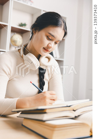 Young Asian woman studying at home office, writing notes with books, laptop and headphones on desk. Online learning, remote education, exam preparation, focus and productivity concept. Young Asian woman studying at home office, writing notes with books, laptop and headphones on desk. Online learning, remote education, exam preparation, focus and productivity concept. 139373410