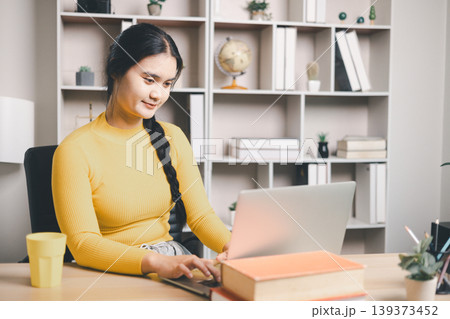 Young Asian woman using laptop at home office desk with books and coffee, studying online or working remotely. Smiling student in modern workspace, e-learning, productivity and lifestyle concept. 139373452