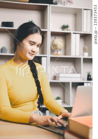 Young Asian woman using laptop at home office desk with books and coffee, studying online or working remotely. Smiling student in modern workspace, e-learning, productivity and lifestyle concept. 139373453