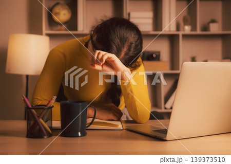 Exhausted young Asian woman covering eyes while sitting by laptop in cozy home office at night, illustrating burnout, study stress, work fatigue, deadline pressure, and late evening overload. Exhausted young Asian woman covering eyes while sitting by laptop in cozy home office at night, illustrating burnout, study stress, work fatigue, deadline pressure, and late evening overload. 139373510