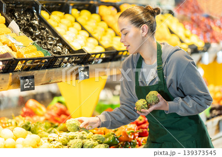 Young woman seller puts artichokes in vegetable shop Young woman seller puts artichokes in vegetable shop 139373545