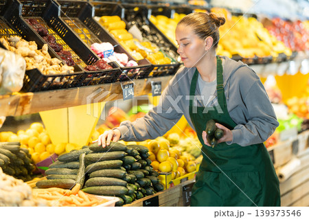 saleswoman stands near a display case and checks the quality of cucumbers saleswoman stands near a display case and checks the quality of cucumbers 139373546