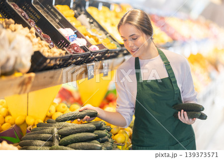 Female seller checks quality of cucumbers and lays them out on counter in supermarket Female seller checks quality of cucumbers and lays them out on counter in supermarket 139373571
