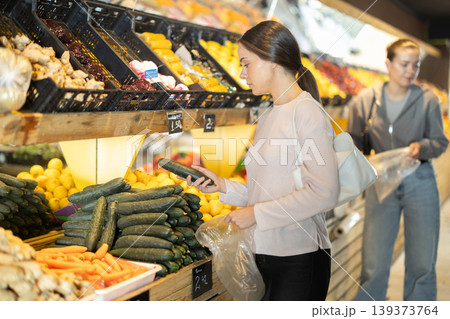 Woman chooses vegetables in a supermarket, she puts fresh cucumbers in a bag Woman chooses vegetables in a supermarket, she puts fresh cucumbers in a bag 139373764